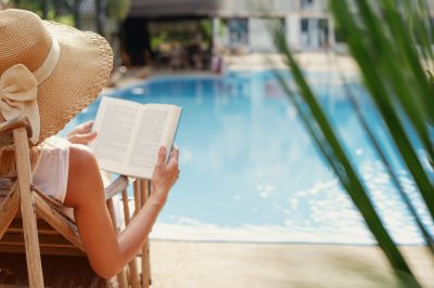 Woman wearing a hat reading a book by a pool